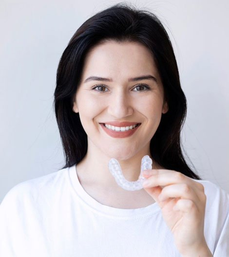 Young woman smiling while holding clear aligner, wearing white shirt against neutral background.