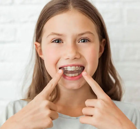 Young girl pointing to her smile against white brick background, displaying healthy teeth.