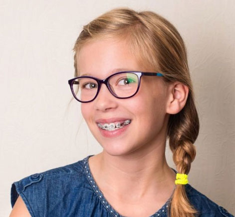 Young girl wearing glasses and denim shirt smiling at camera in dental office setting.