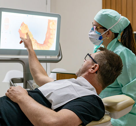 Dentist in green scrubs and mask shows male patient X-ray results on digital monitor during dental consultation.