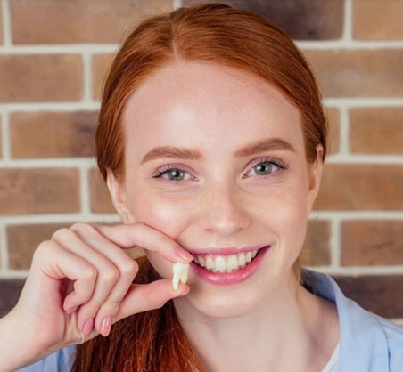 Red-haired woman smiling brightly against brick wall, touching teeth with finger.