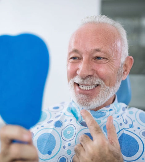 Smiling senior man in blue patterned shirt holding dental mirror in modern dental office setting.