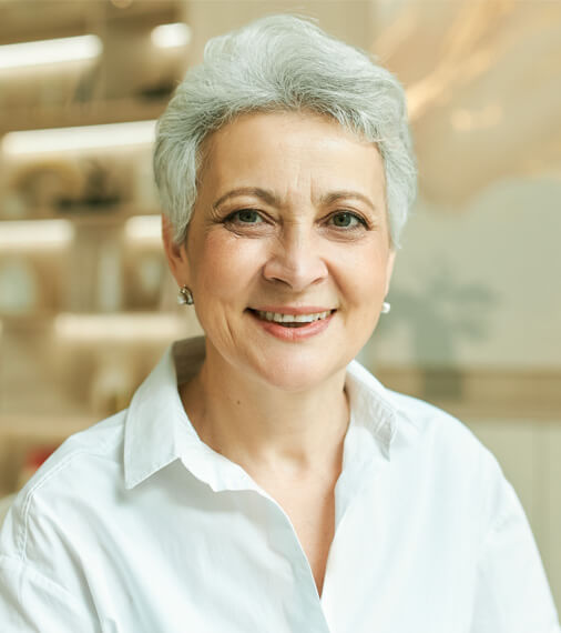 Smiling older woman with gray hair wearing white shirt in bright dental office setting.