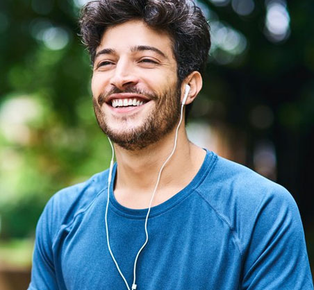 Young man smiling outdoors wearing blue shirt and earbuds, green blurred background.