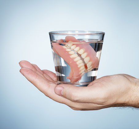 Hand holding glass of water with dentures submerged inside against blue background.