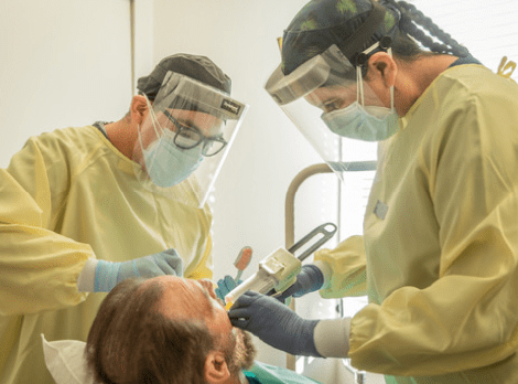 Two dental professionals in yellow protective gear examine a patient's teeth in a clinical setting.